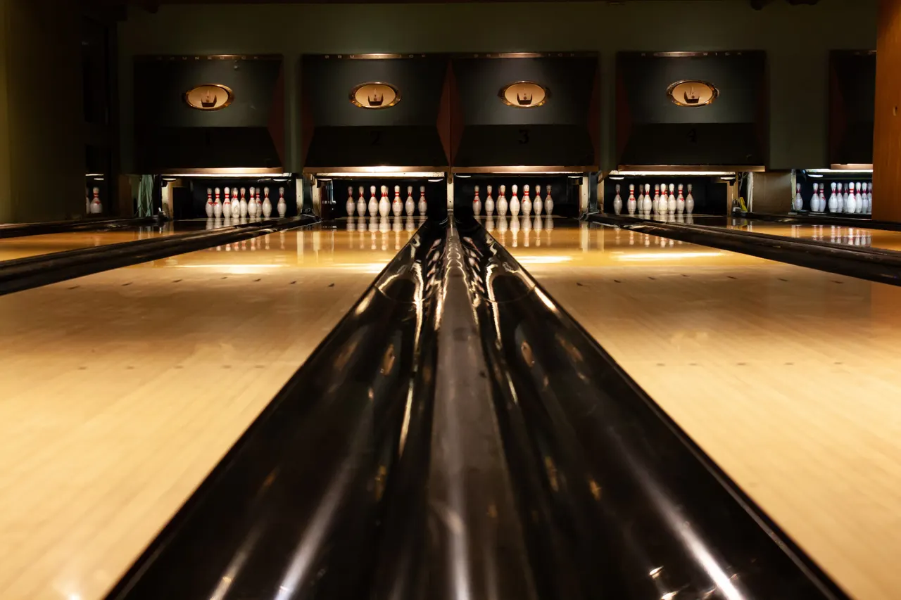 Multiple empty bowling lanes with white pins set up, viewed along the polished wooden floor under warm lighting.