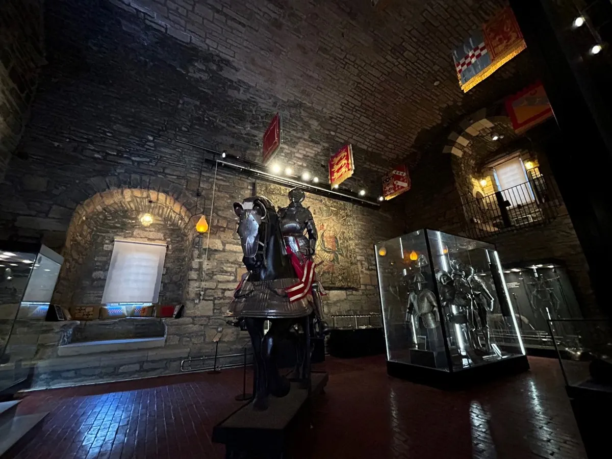 Stone castle interior with medieval armour displays and glass exhibition cases lit by spotlights at night.