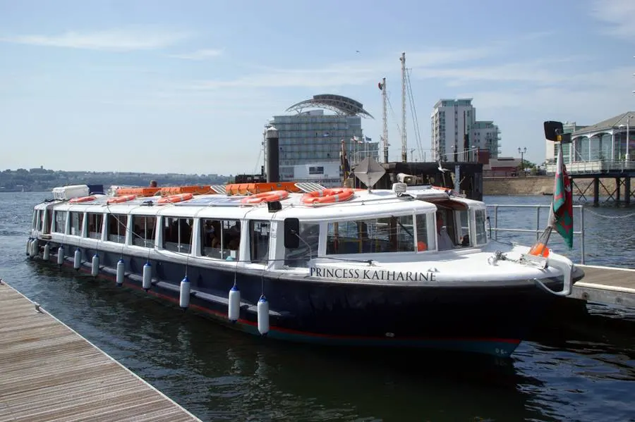Passenger riverboat moored at a wooden pier on the water, with modern buildings visible across the river.