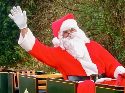 A person dressed as Father Christmas waves from a decorated railway carriage with evergreen foliage in the background.