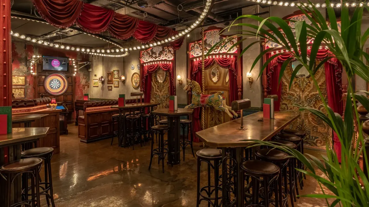 Ornate vintage-style bar interior with red velvet drapes, brass lighting, wooden tables and stools, and decorative wall panels.
