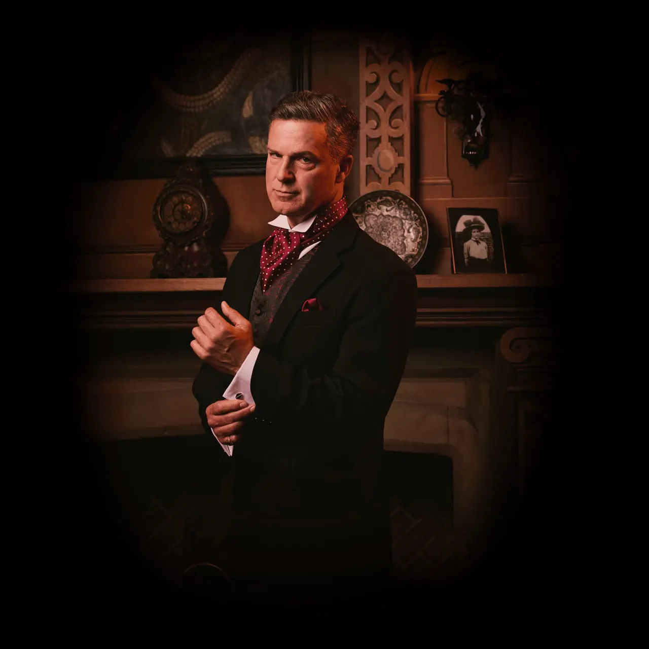 Man in formal black suit and red polka-dot bow tie posing in front of ornate antique decor and period furnishings.