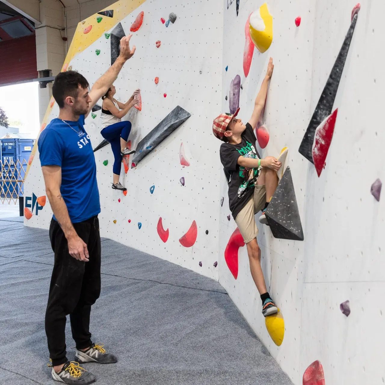 A man in a blue shirt watches a young boy climb an indoor wall with colorful handholds.