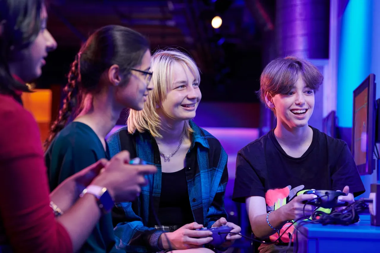 Young people playing video games together under purple and blue lighting in an arcade or gaming space.
