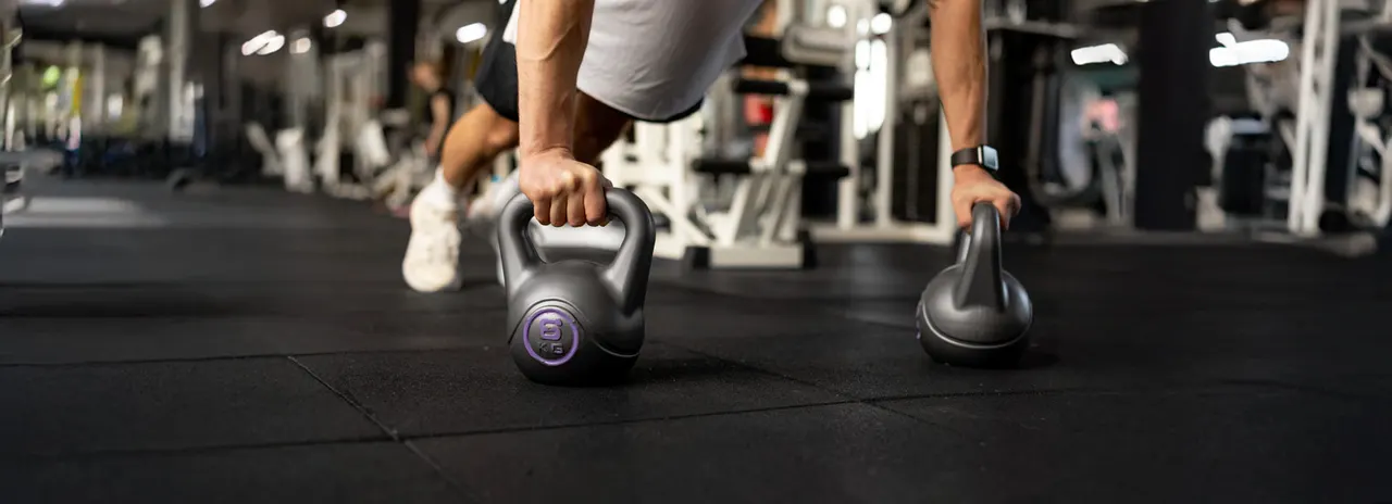 Person performing kettlebell exercise on gym floor with fitness equipment in background.