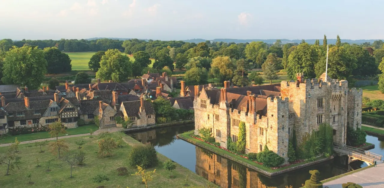 Aerial view of a medieval stone castle surrounded by a moat, with Tudor buildings and manicured grounds, set within a landscape of woodland and fields.