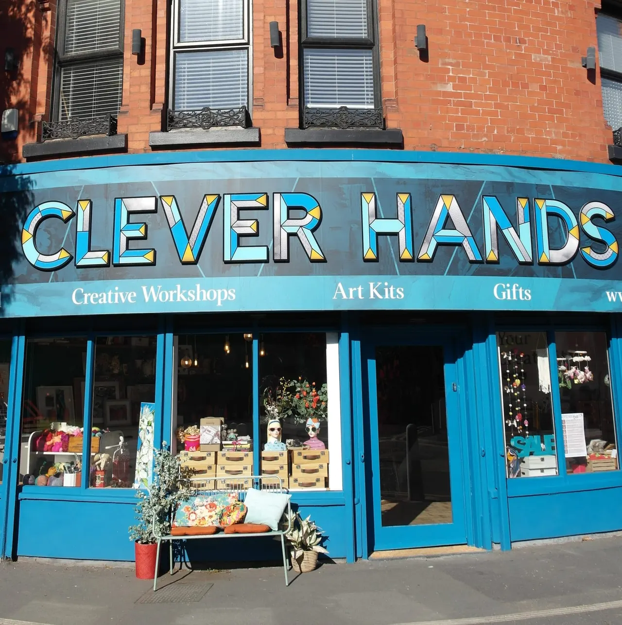 Bright blue storefront with large signage, displaying art kits and gifts in windows, with potted plants outside on the pavement.