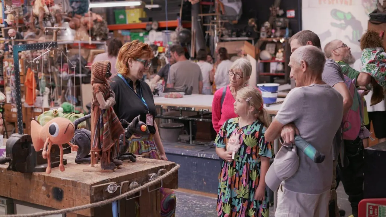 Visitors of various ages gather around a puppet display demonstration at an indoor venue, with a staff member presenting puppets to the audience.