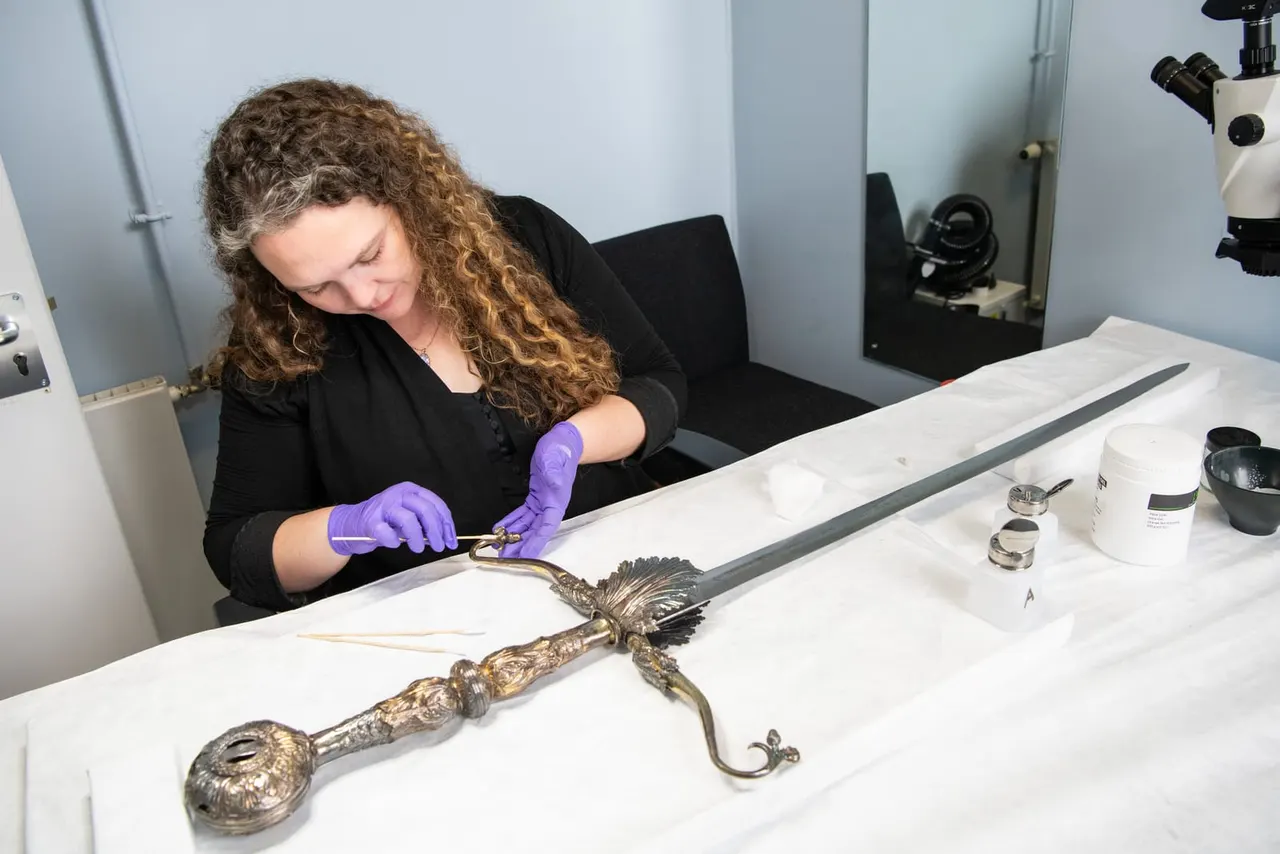 This is a PHOTOGRAPH.

Conservator in purple gloves cleaning an ornate historical bronze sword or blade at a laboratory workbench.