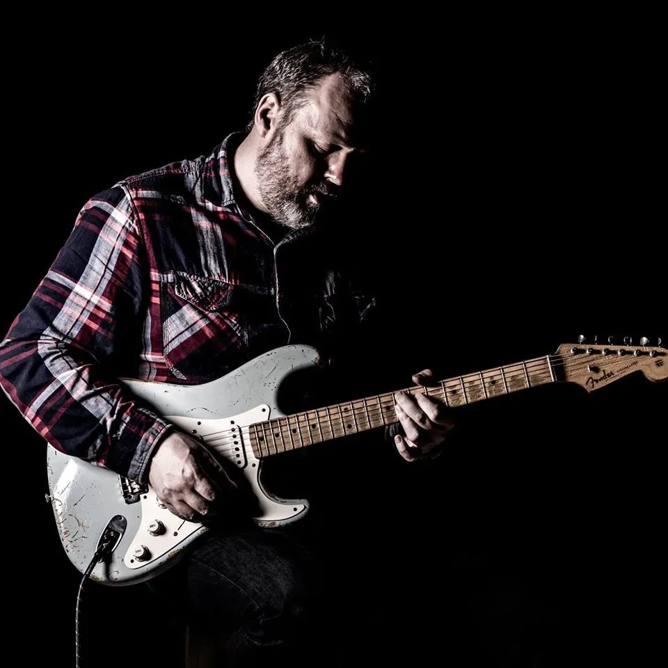 Man playing electric guitar against a black background, wearing a red and navy checked shirt.