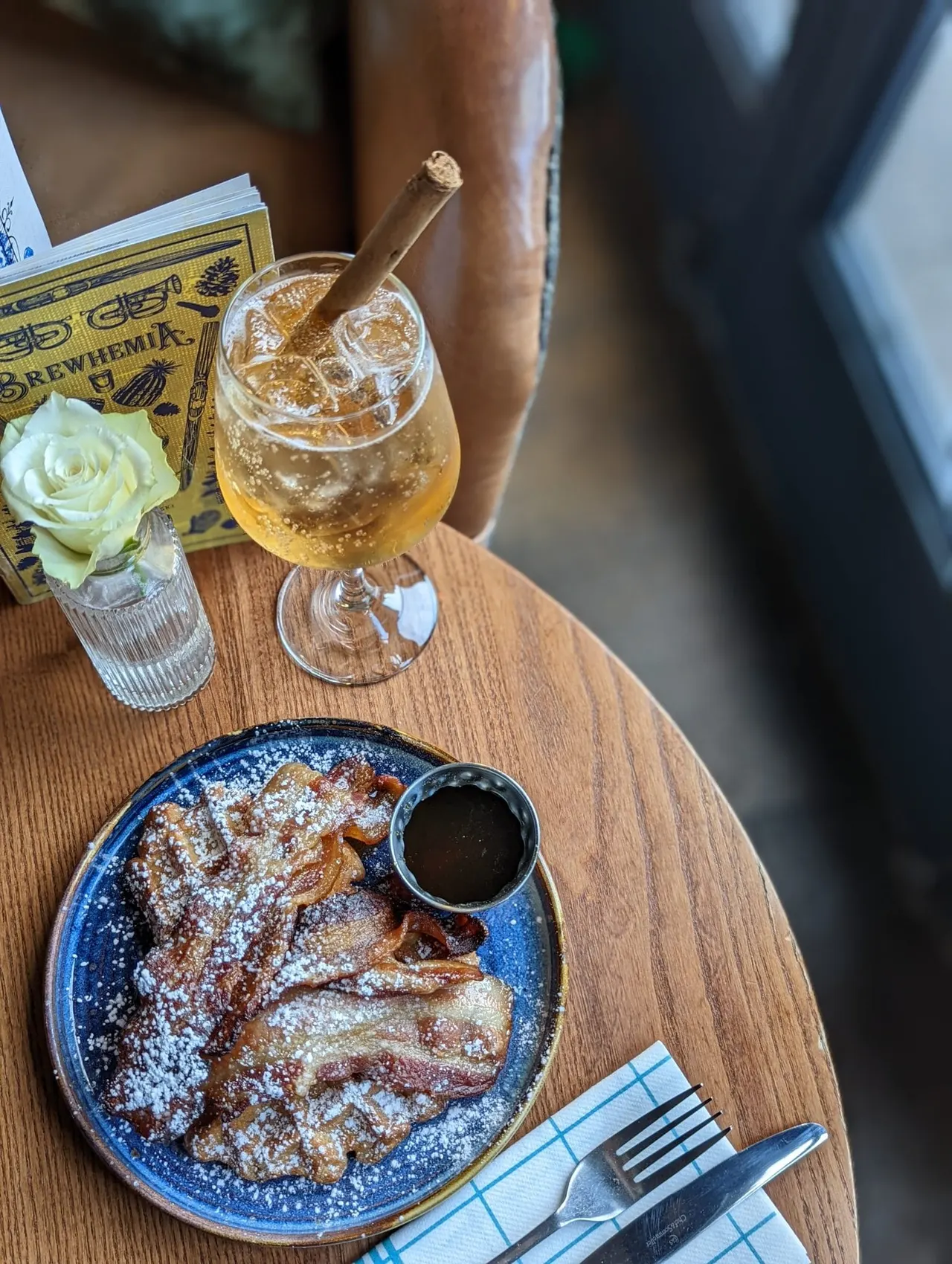 Person holding a cocktail with a cigar beside a wooden table displaying pastries, a drink, and dining utensils.
