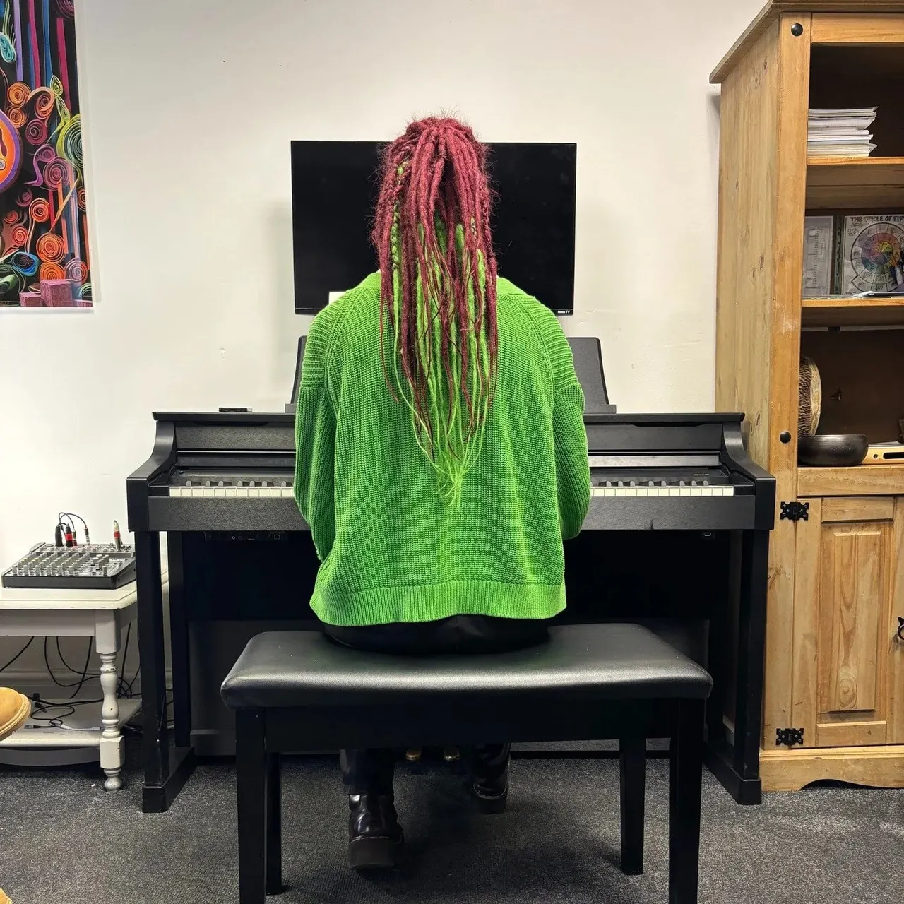 Person with long pink and green dreadlocks wearing a bright green jumper, seated at a black digital piano in a music studio.