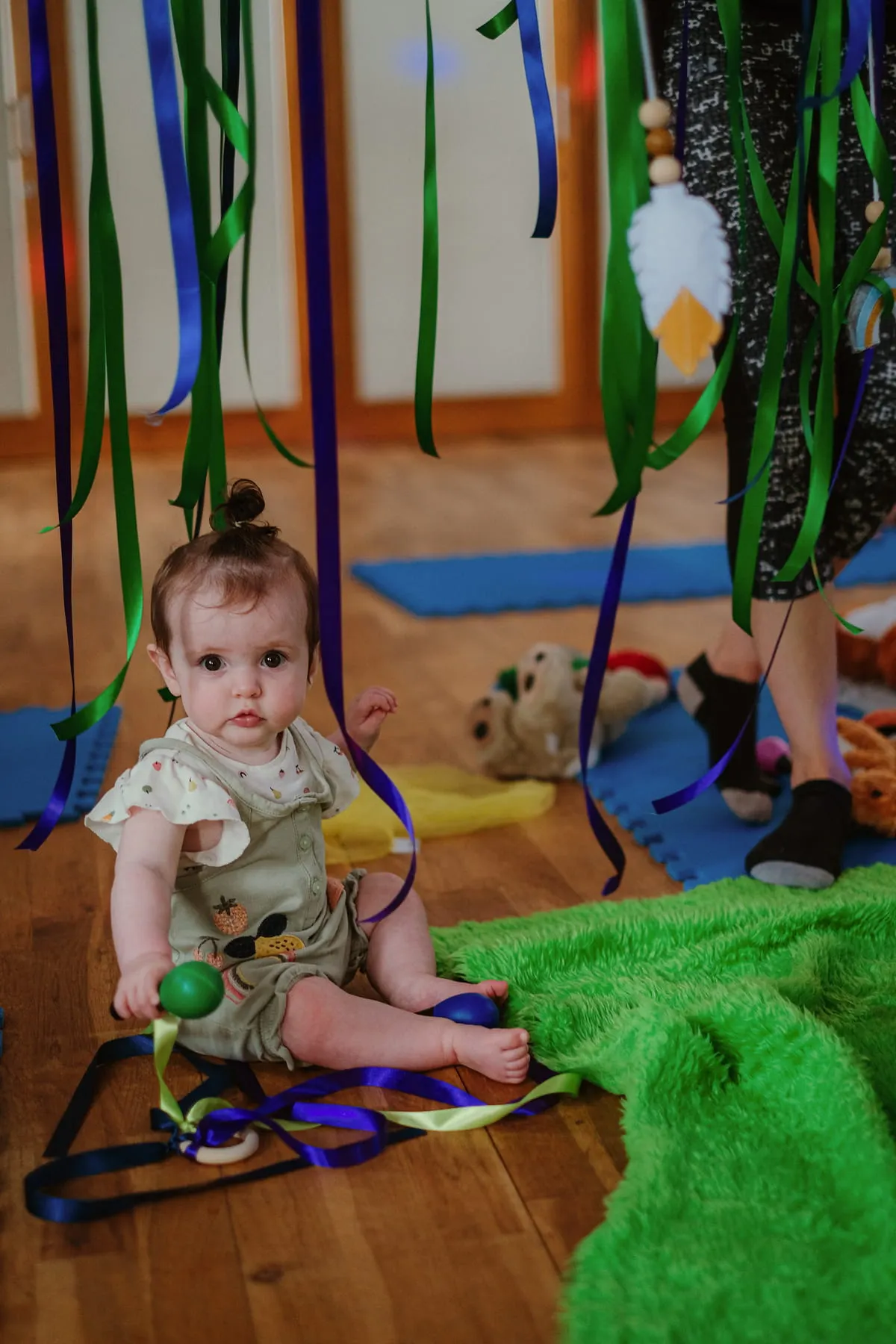 A toddler in a beige outfit sits on a wooden floor surrounded by coloured ribbons and green textured play mats in an indoor play space.