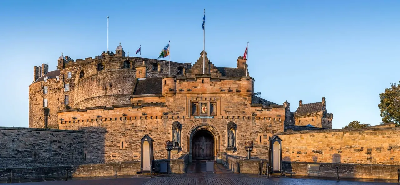 This is a PHOTOGRAPH.

Edinburgh Castle's stone fortress with multiple turrets and flags flying, viewed from the entrance gatehouse under clear blue sky.