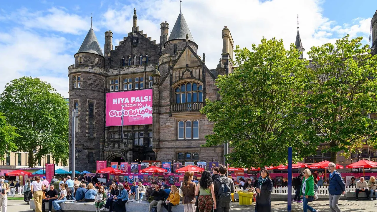 Outdoor event with crowds gathered under red umbrellas in front of a stone castle building with pink banner signage.