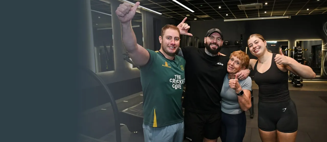 Five people of mixed ages posing together in a gym, giving thumbs up gestures.
