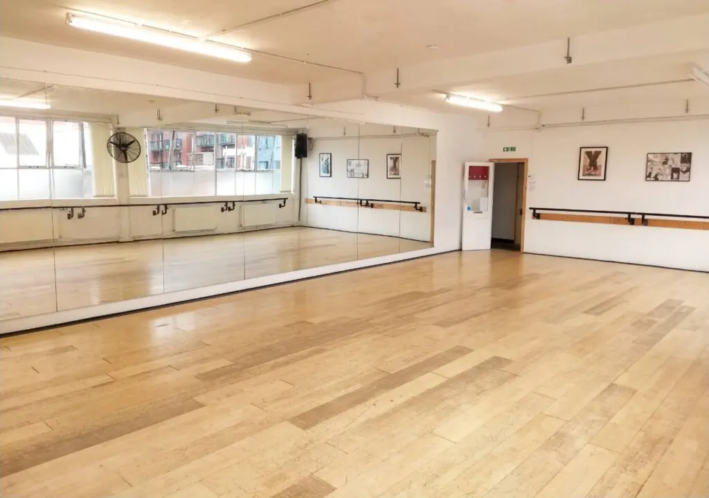 Empty dance studio with wooden floor, mirrored walls, ballet barres, and large windows overlooking a street.