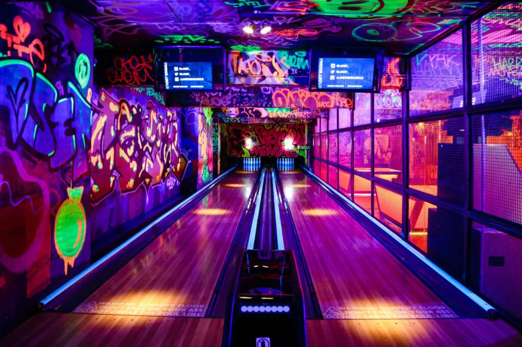 A bowling alley with neon-lit lanes, colourful graffiti-style wall art, and glowing pins at the far end.
