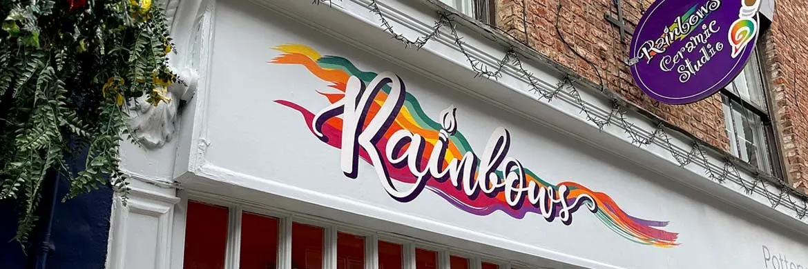 Exterior storefront of Rainbows Ceramics Painting Studio showing colourful logo signage and red doors on a brick building.