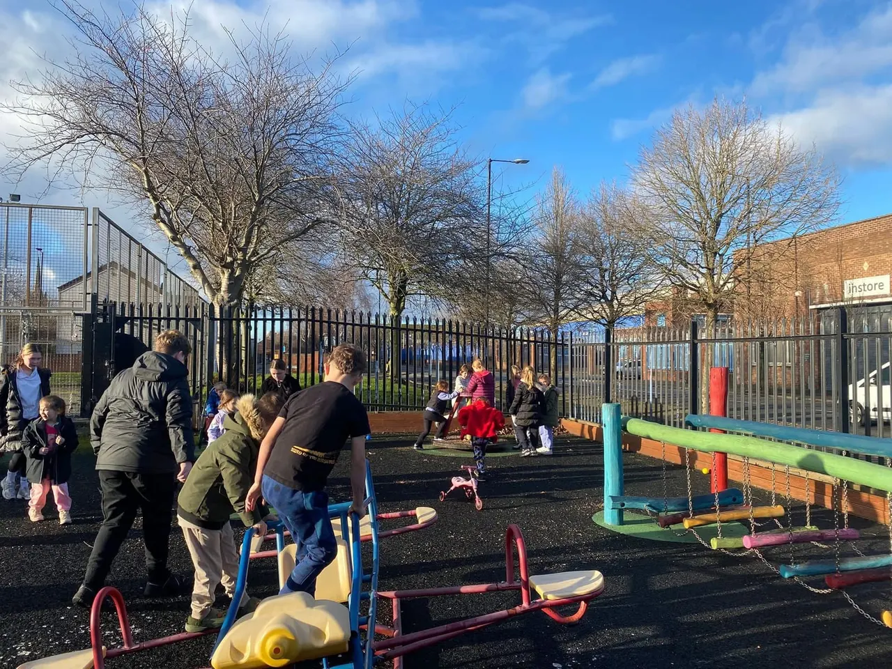 Children and adults playing on colourful climbing equipment and swings in a fenced outdoor play area on a sunny day.