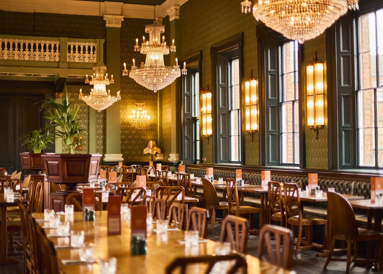 Grand dining room with crystal chandeliers, wooden chairs, and elegant green wallpapered walls lit by wall sconces and tall windows.