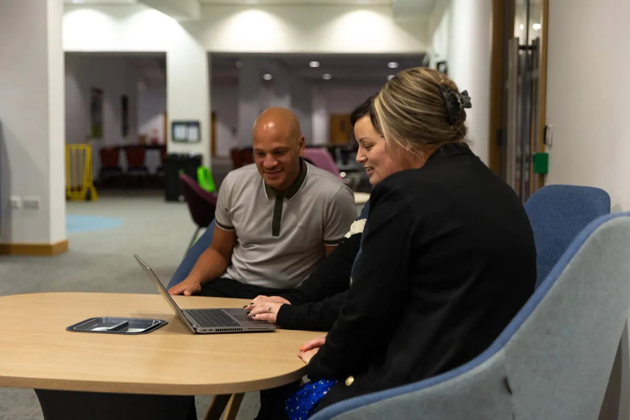 Two people seated at a wooden table in a modern leisure facility, looking at a laptop together.