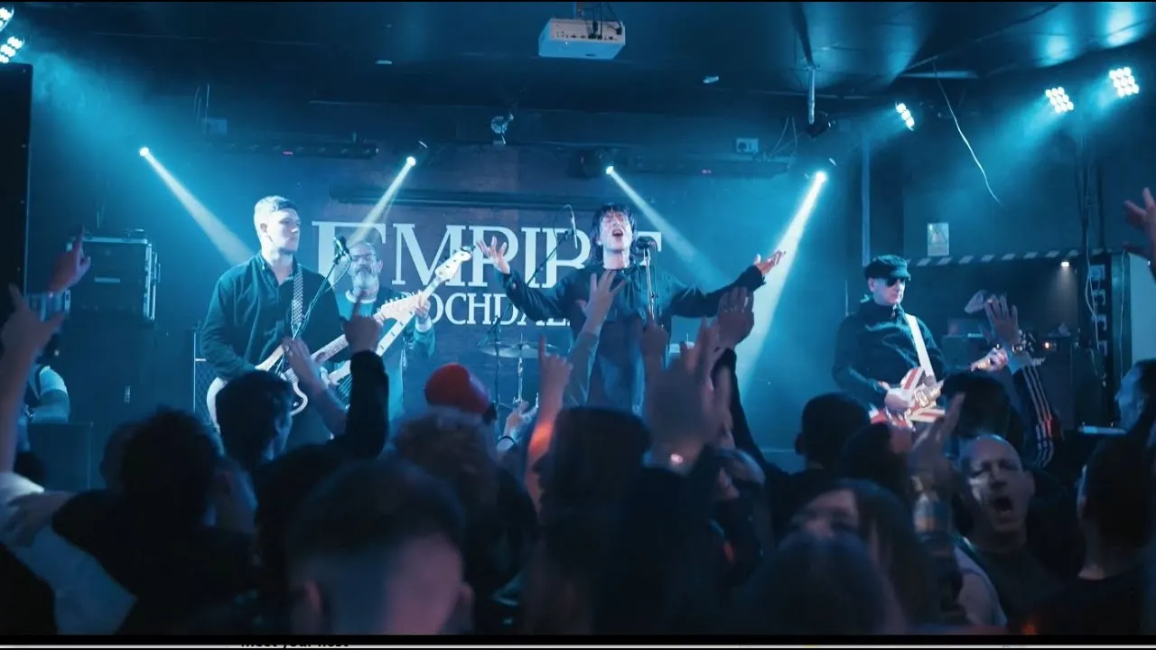 A band performs on stage under blue lighting while an audience watches and raises their hands at an indoor music venue.