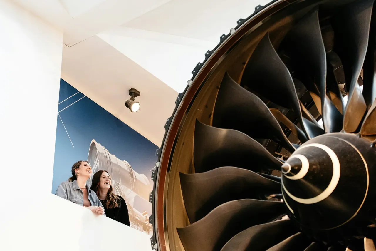 Two visitors look up at a large steam engine propeller on display in a museum gallery.