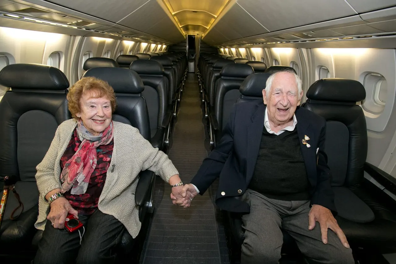Two older adults sitting together in an aircraft cabin holding hands, smiling at the camera.