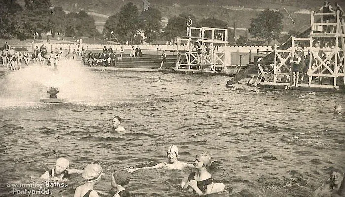 Vintage sepia photograph of swimmers in a large outdoor pool with wooden structures and spectators along the shore.