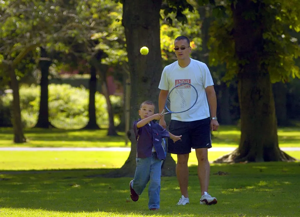 Adult and young child playing tennis together on a sunny park lawn lined with tall trees.