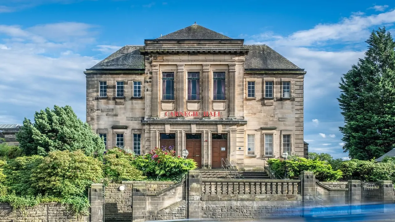 Classical stone theatre building with pitched roof and columned entrance, surrounded by green hedges and gardens under blue sky.
