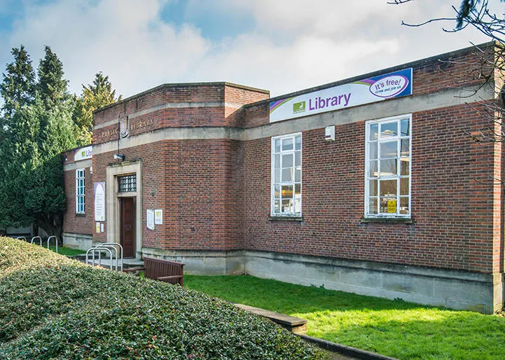 Brick library building with blue signage, surrounded by hedges and grass on a sunny day.