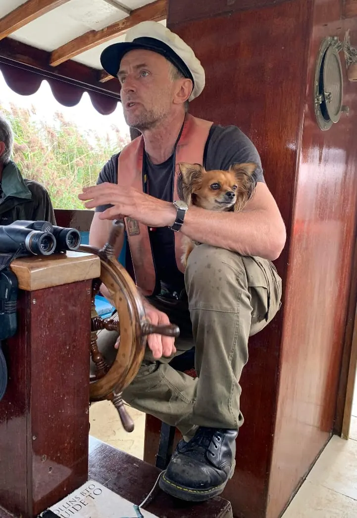Man holding a small dog inside a wooden boat cabin, gesturing whilst giving a commentary during a wildlife tour.
