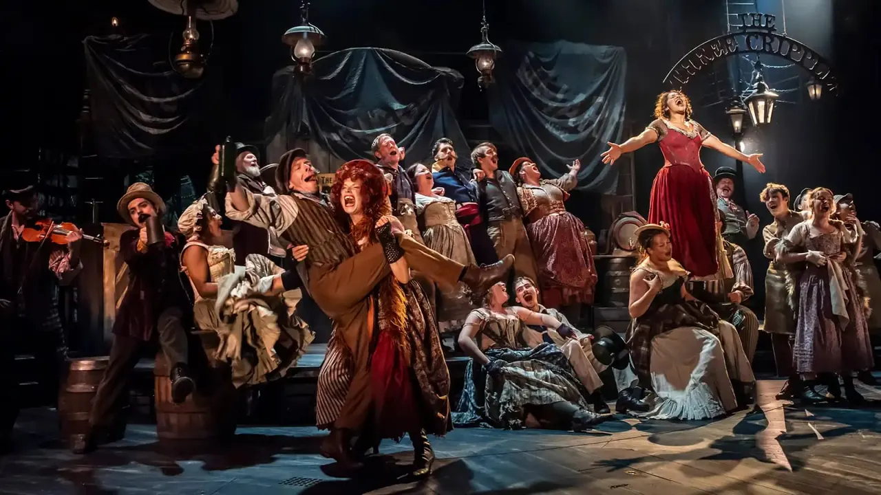 Large theatrical cast performing on stage in period costume with dramatic lighting and dark draped backdrop.