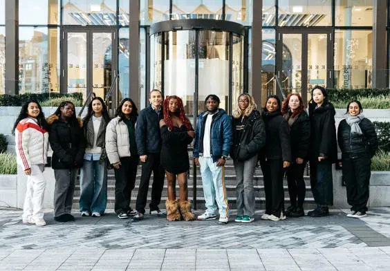 A group of approximately fifteen people standing together outside a modern building with large glass windows and urban backdrop.