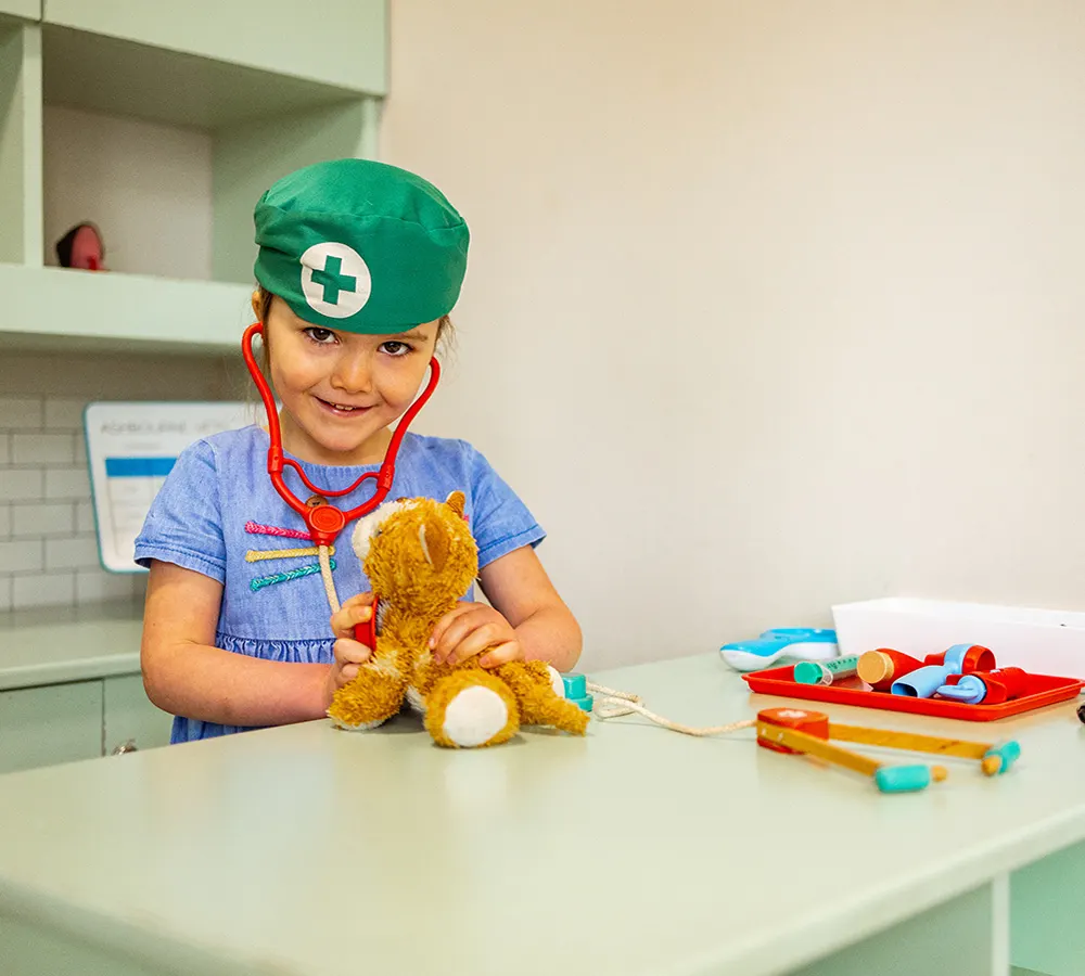 Child wearing medical costume examining a toy bear with stethoscope at a play medical station.