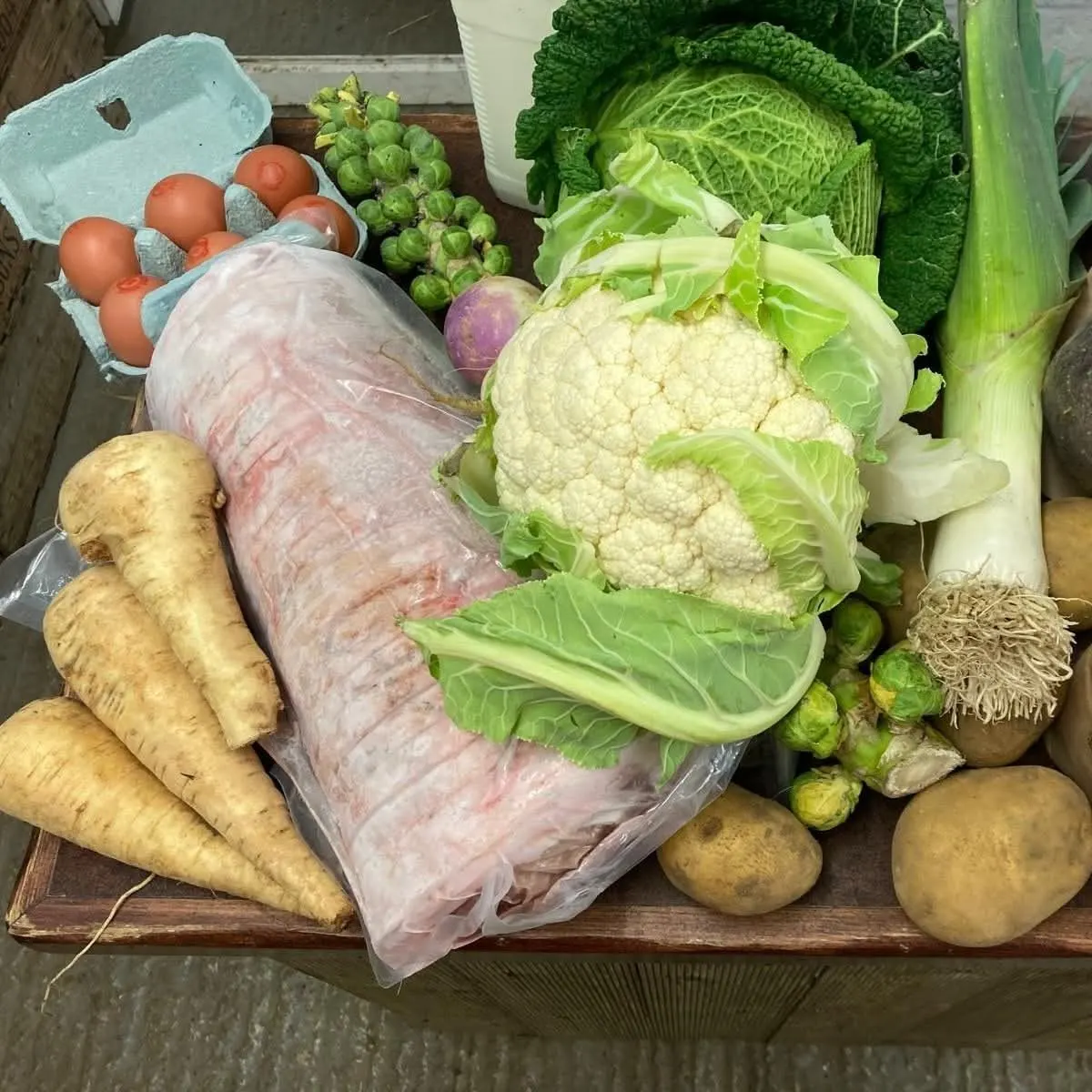 Farm produce display including fresh vegetables, eggs, and wrapped meat on a wooden board.