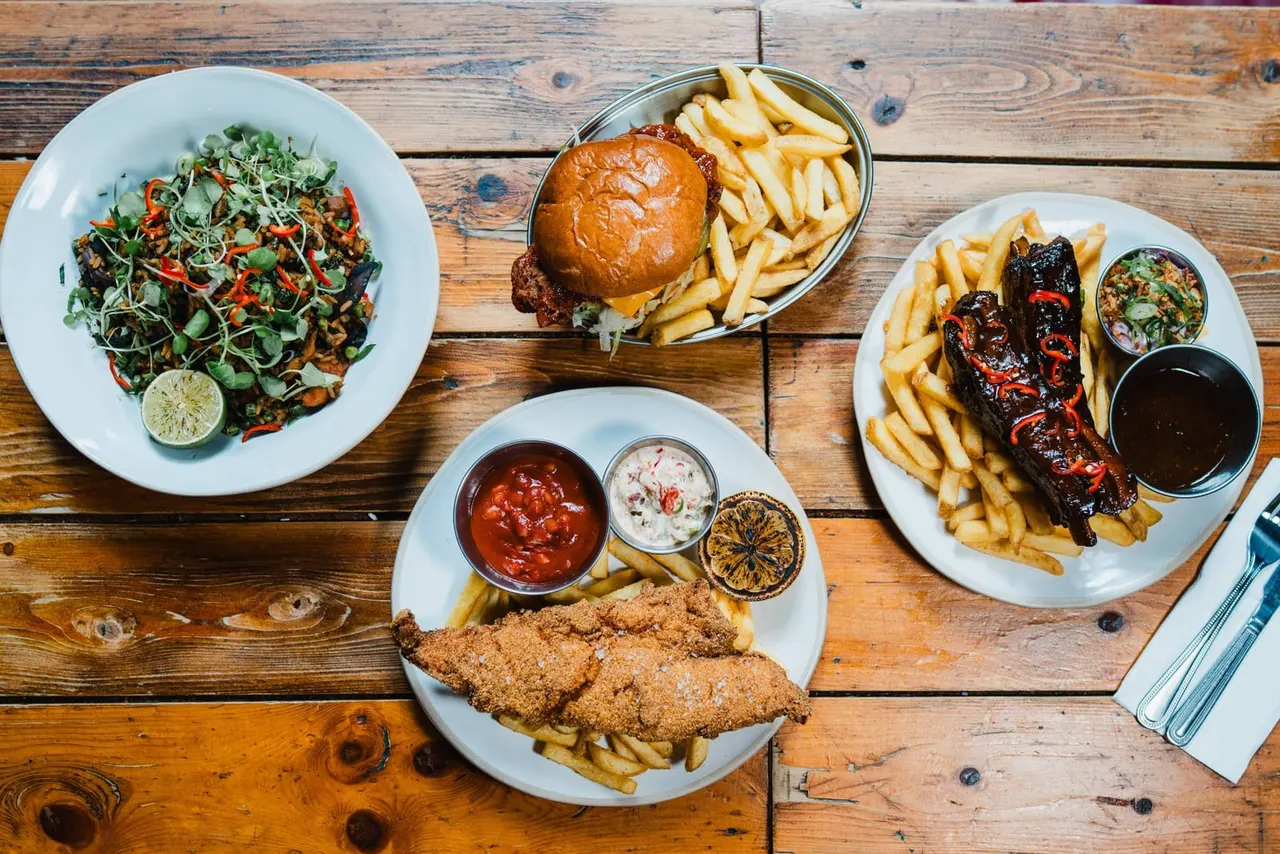 Overhead view of four restaurant dishes on a wooden table: green salad with microgreens, burger with fries, fried chicken with fries and sauces, and glazed ribs with fries.