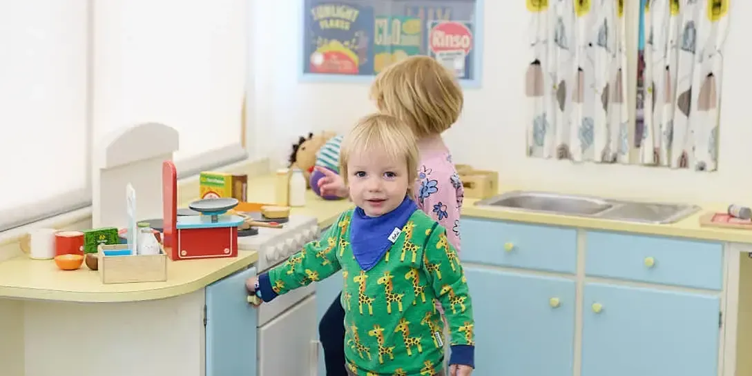 A toddler in a green patterned jumper plays at a toy kitchen whilst an adult supervises in the background.
