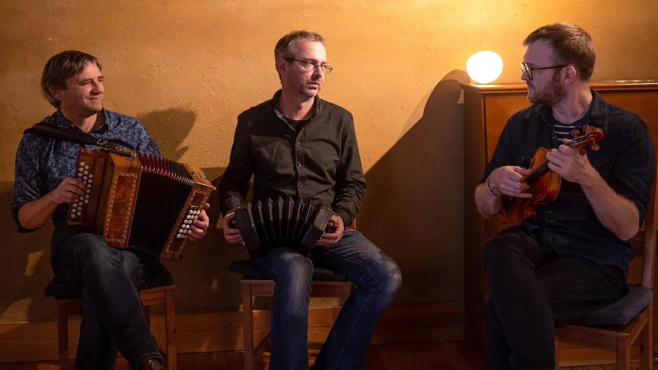 Three musicians seated indoors playing accordion and fiddles against a warm ochre-coloured wall.