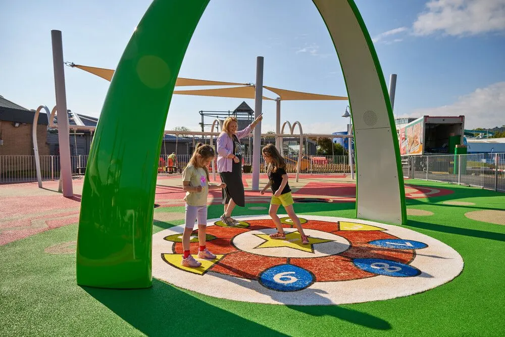 Children playing on a colourful hopscotch game under a large green arch structure at an outdoor leisure park.