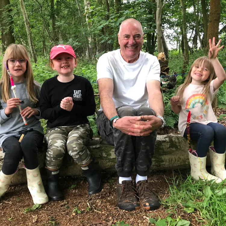 An adult and three children sit together on a log in a woodland setting, the adult holding something in his hand whilst smiling at the camera.