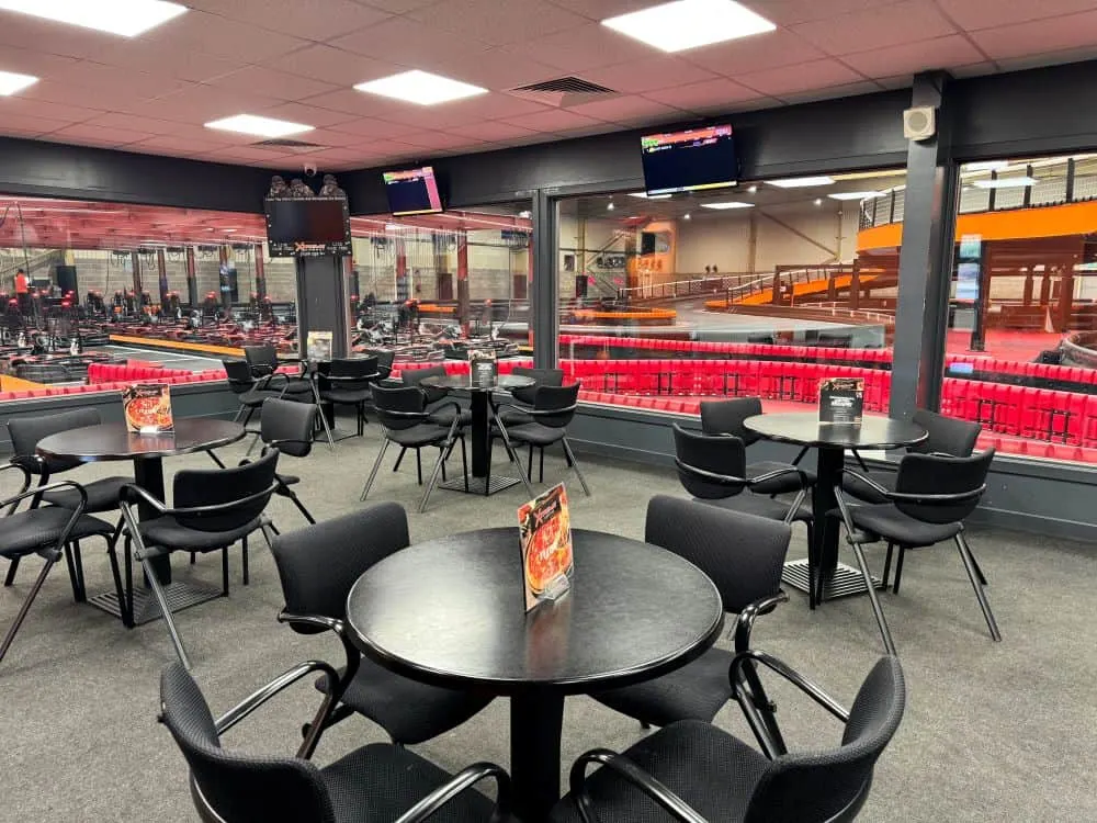 Indoor seating area overlooking a go-kart racing track with red barriers and orange kart, multiple round tables with black chairs.