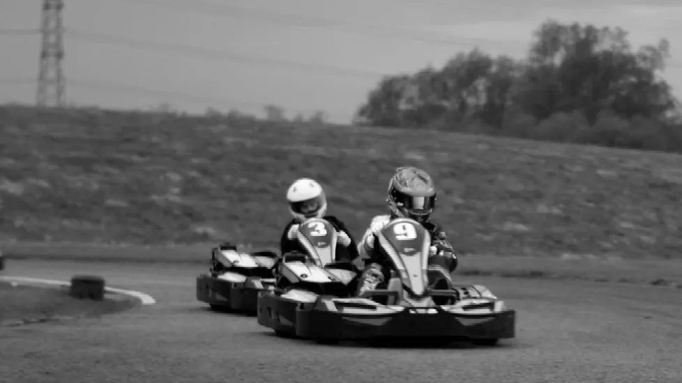 Two go-kart drivers racing on an outdoor track, wearing helmets and numbered vehicles.