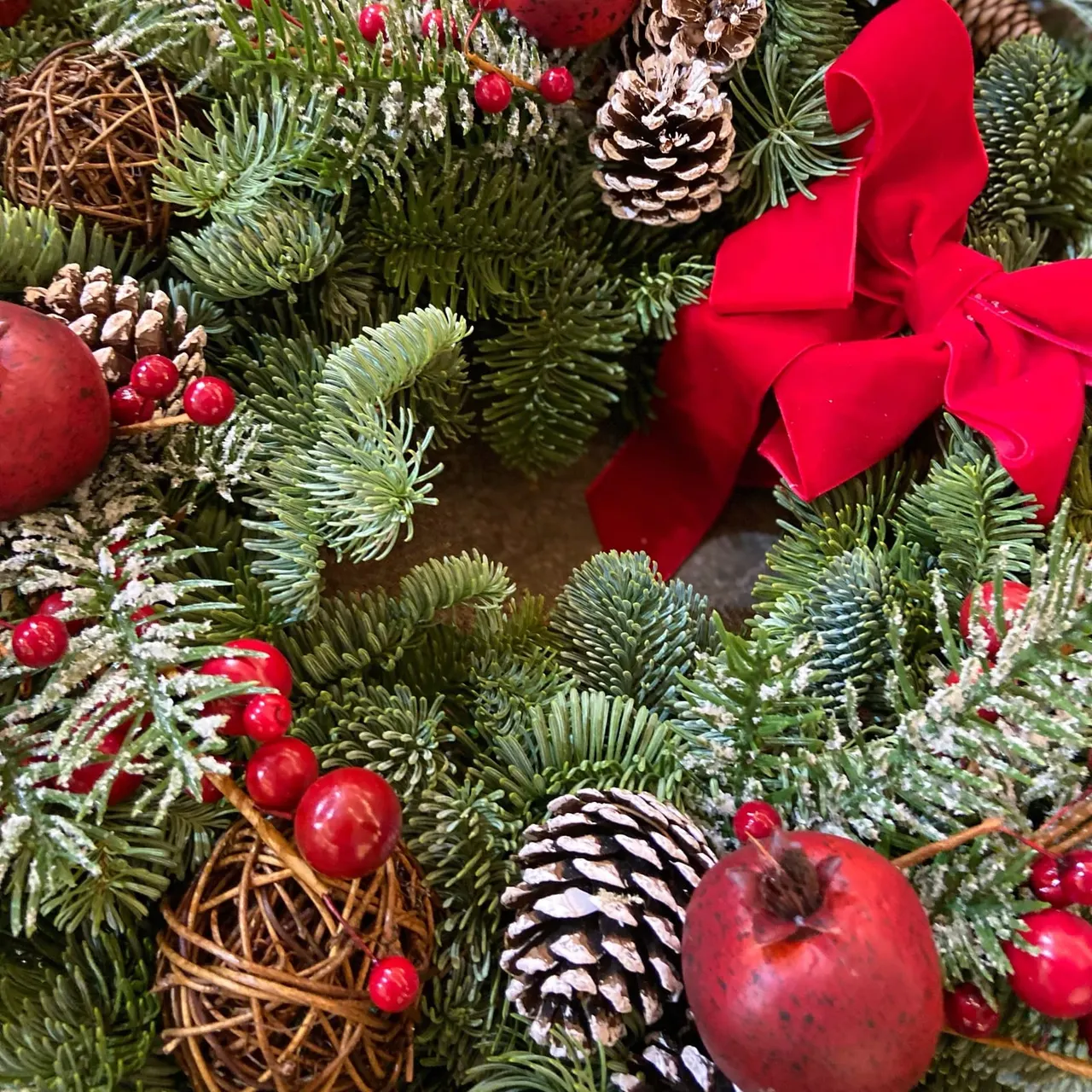 Close-up of festive Christmas wreath decorated with evergreen branches, red baubles, berries, pinecones, wicker spheres and a bold red ribbon bow.