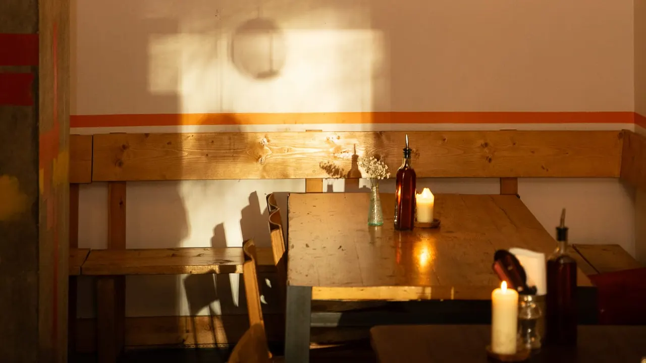 Warm sunlight floods a wooden dining table set with bottles, candles and flowers in a rustic restaurant interior.