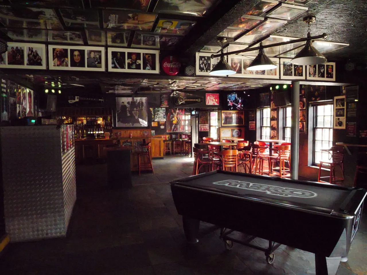 Interior of a bar and leisure venue with pool table, red seating area, bar counter, and walls decorated with photographs and memorabilia.
