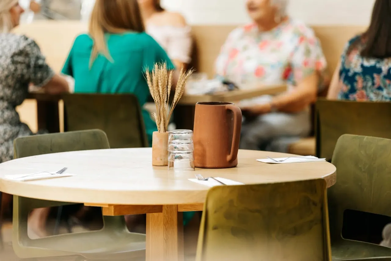 Round wooden table with ceramic jug, glass and dried flowers, blurred group of people seated in background.