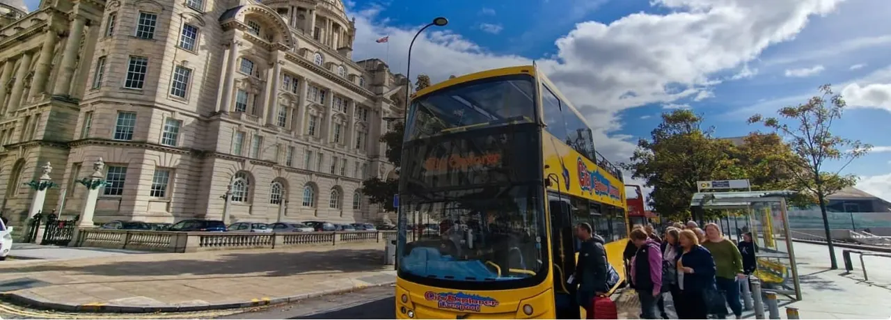 Double-decker yellow tour bus with passengers boarding beside a grand stone building and bus shelter on a sunny day.